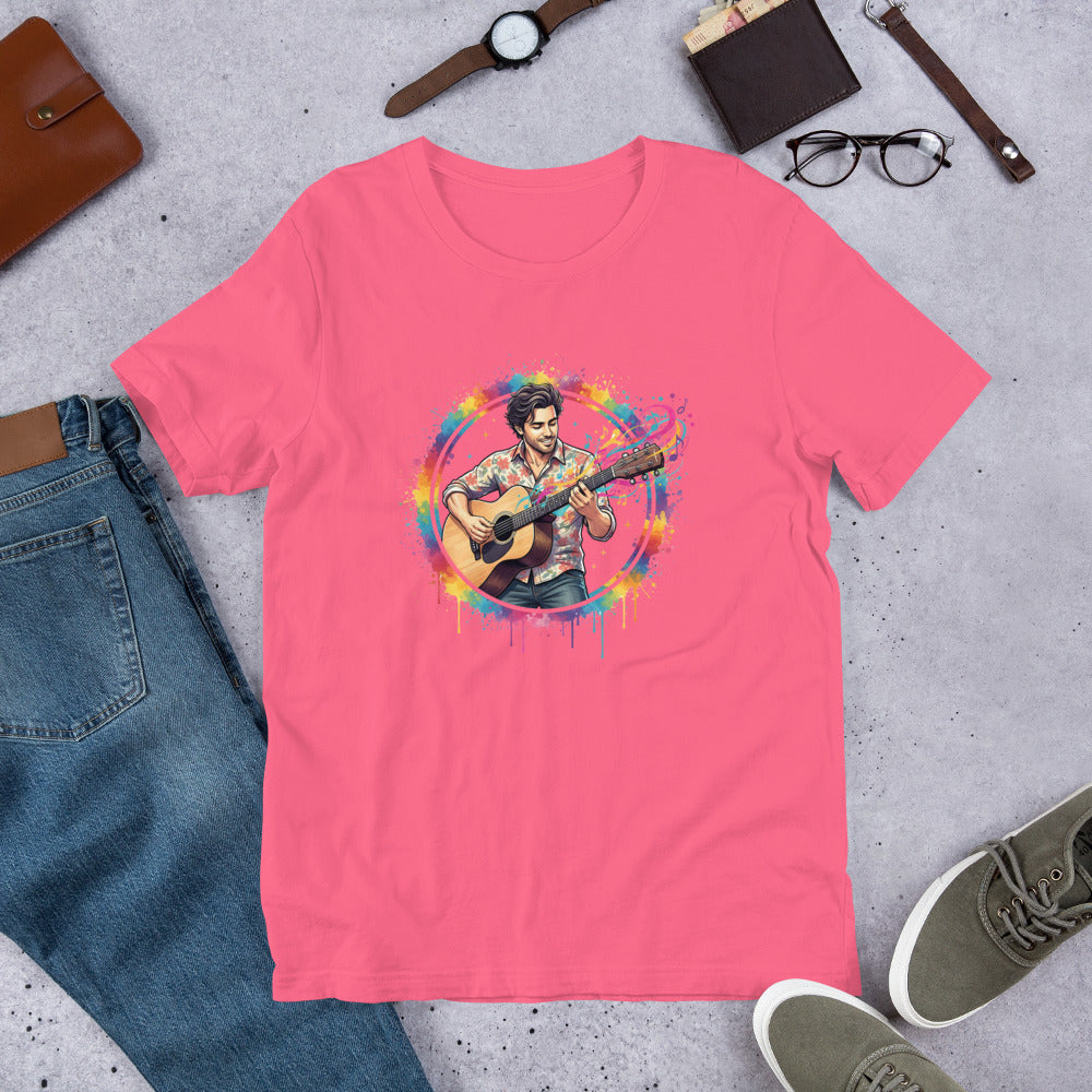 Pink t-shirt with a colorful graphic of a person playing guitar, surrounded by jeans, shoes, and accessories on a concrete surface.